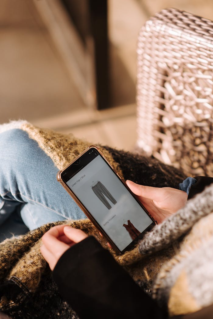 High angle of unrecognizable woman sitting on chair and choosing clothes in internet shop using mobile phone